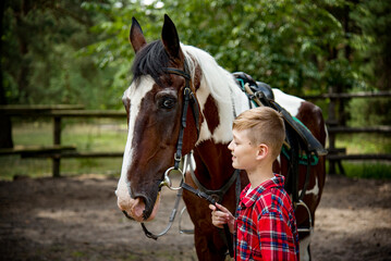 Obraz premium little handsome blonde smiling boy in red checkered shirt hugging horse in green forest on sunny day 