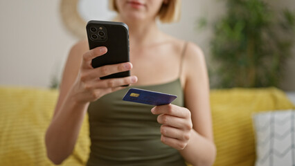 Young blonde woman shopping with smartphone and credit card sitting on sofa at home