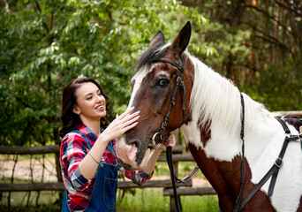 Obraz premium portrait of beautiful smiling brunette cowboy woman in red checkered shirt walking and hugging with horse in green forest on sunny day