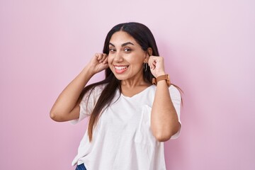 Young arab woman standing over pink background smiling pulling ears with fingers, funny gesture....