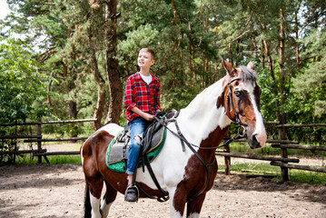 little handsome blonde smiling  boy  in red checkered shirt riding  horse in green forest on sunny day 
