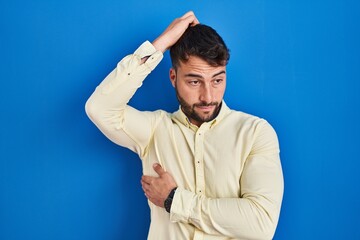 Handsome hispanic man standing over blue background confuse and wondering about question. uncertain with doubt, thinking with hand on head. pensive concept.