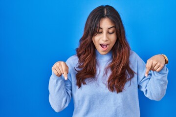 Fototapeta premium Hispanic young woman standing over blue background pointing down with fingers showing advertisement, surprised face and open mouth