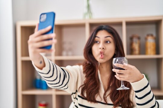 Hispanic young business woman taking a selfie picture drinking a glass of wine making fish face with mouth and squinting eyes, crazy and comical.
