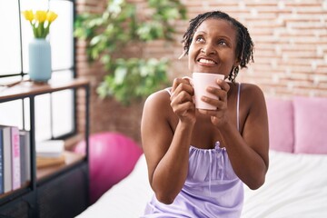 Middle age african american woman drinking cup of coffee sitting on bed at bedroom