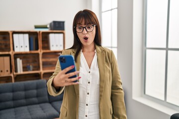 Young brunette woman working at the office with smartphone scared and amazed with open mouth for surprise, disbelief face