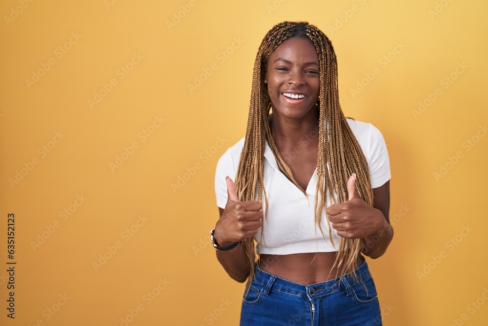 Sticker African american woman with braided hair standing over yellow background success sign doing positive gesture with hand, thumbs up smiling and happy. cheerful expression and winner gesture.