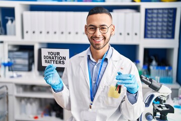 Young hispanic man working at scientist laboratory holding your donation matters holding blood sample smiling with a happy and cool smile on face. showing teeth.