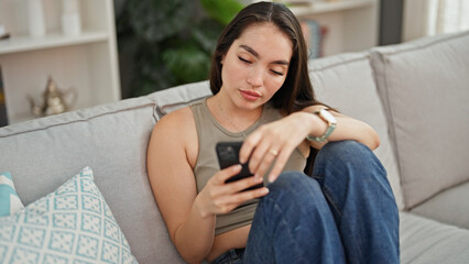 Obraz premium Young beautiful hispanic woman using smartphone sitting on sofa at home
