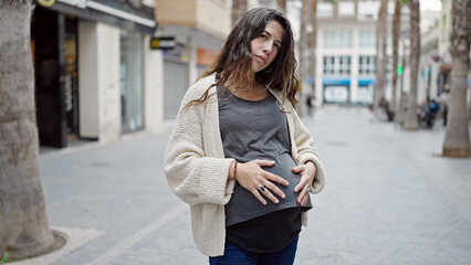 Young pregnant woman standing with serious expression touching belly at street