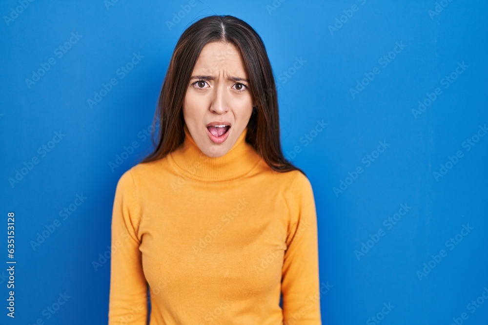 Poster young brunette woman standing over blue background in shock face, looking skeptical and sarcastic, s