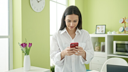 Young beautiful hispanic woman using smartphone standing at dinning room