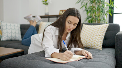 Young beautiful hispanic woman writing on notebook lyin on sofa at home
