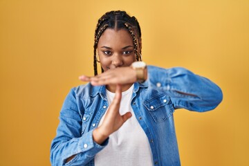 African american woman with braids standing over yellow background doing time out gesture with...