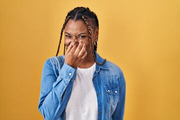 African american woman with braids standing over yellow background smelling something stinky and...