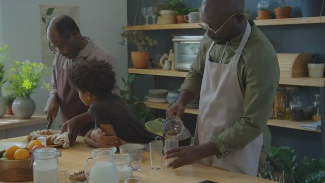 African American man pouring Zobo tea into glasses while his senior father and little son cutting Meskouta cake in kitchen at home