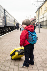 small baby boy stands on the platform near the train and holds a yellow suitcase in his hands.