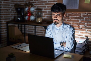 Young hispanic man with beard working at the office at night skeptic and nervous, disapproving...