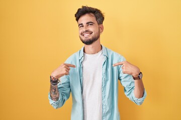Young hispanic man with tattoos standing over yellow background looking confident with smile on...