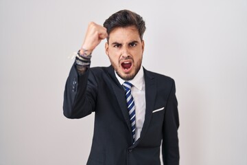 Young hispanic man with tattoos wearing business suit and tie angry and mad raising fist frustrated and furious while shouting with anger. rage and aggressive concept.