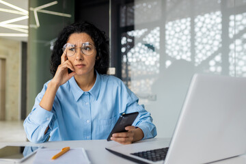 Sad upset business woman at workplace, Hispanic woman disappointed with work results inside office, female worker using laptop, holding smartphone.