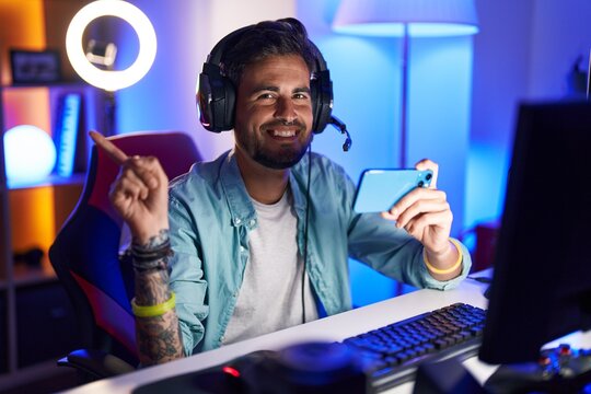 Young hispanic man with tattoos playing video games with smartphone smiling happy pointing with hand and finger to the side