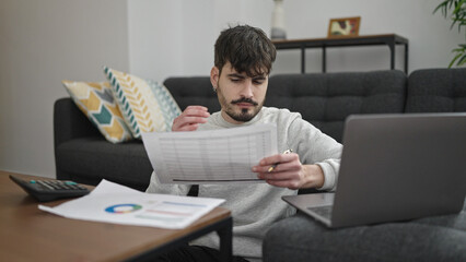 Young hispanic man using laptop sitting on sofa working at home