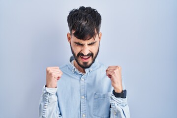 Young hispanic man with beard standing over blue background excited for success with arms raised and eyes closed celebrating victory smiling. winner concept.