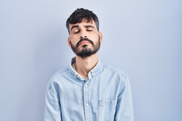 Young hispanic man with beard standing over blue background looking sleepy and tired, exhausted for...