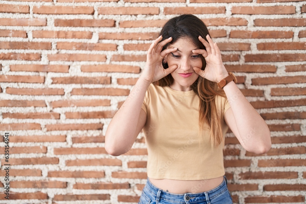 Sticker young brunette woman standing over bricks wall trying to open eyes with fingers, sleepy and tired fo