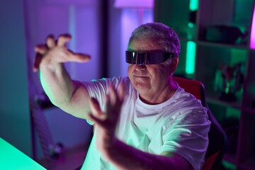 Middle age grey-haired man streamer playing video game using virtual reality glasses at gaming room