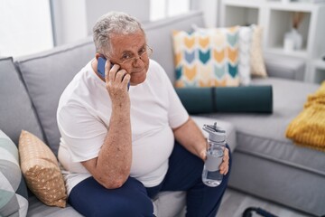 Middle age grey-haired man talking on smartphone holding bottle of water at home