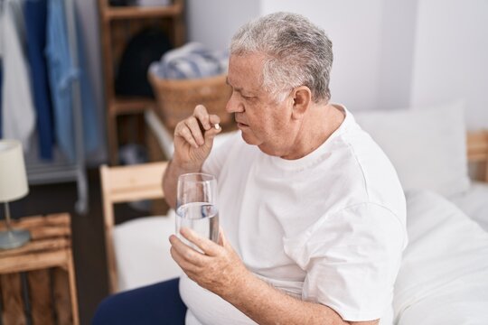 Middle age grey-haired man taking pill drinking water sitting on bed at bedroom