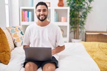 Hispanic young man using computer laptop sitting on the bed looking positive and happy standing and smiling with a confident smile showing teeth