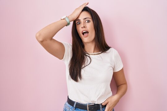Young brunette woman standing over pink background surprised with hand on head for mistake, remember error. forgot, bad memory concept.