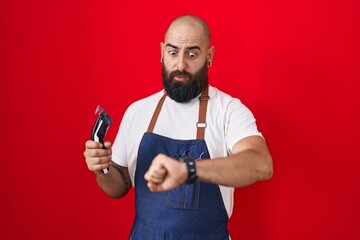 Young hispanic man with beard and tattoos wearing barber apron holding razor looking at the watch time worried, afraid of getting late