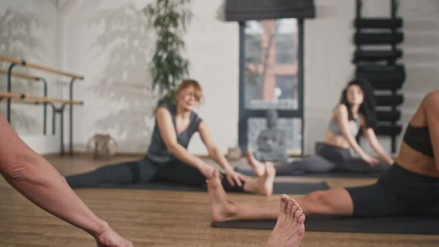 Sporty Caucasian women practicing stretching on yoga mats. Wearing fitness clothes while training in modern studio. Females improving their body flexibility. Concept of meditation.