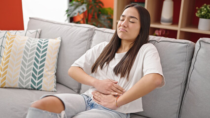 Young beautiful hispanic woman suffering for stomach ache sitting on sofa at home