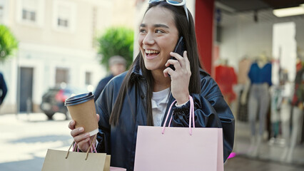 Young beautiful hispanic woman talking on smartphone holding shopping bags and coffee at street