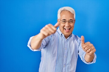 Hispanic senior man wearing glasses approving doing positive gesture with hand, thumbs up smiling and happy for success. winner gesture.