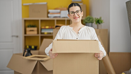 Young beautiful hispanic woman smiling confident holding package at new home