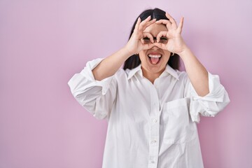Young brunette woman standing over pink background doing ok gesture like binoculars sticking tongue out, eyes looking through fingers. crazy expression.