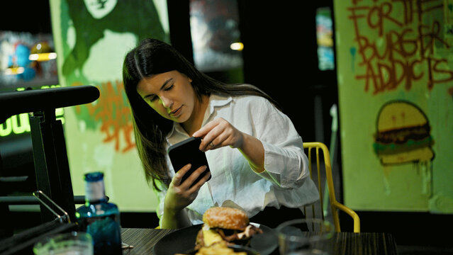 Young beautiful hispanic woman taking picture of delicious hamburger with smartphone at the restaurant