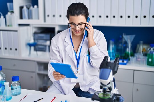 Young hispanic woman scientist talking on smartphone using touchpad at laboratory