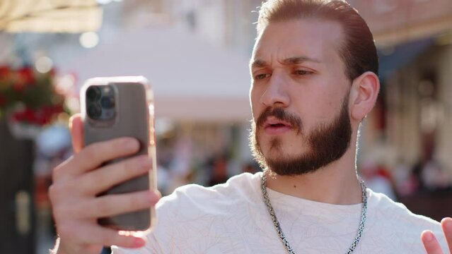 Oh my God Wow. Excited happy joyful winner bearded young man use smartphone typing browsing celebrating win good message news. Guy with mobile phone walking in urban sunshine city street outdoors