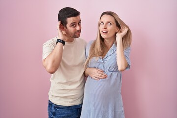 Young couple expecting a baby standing over pink background smiling with hand over ear listening an hearing to rumor or gossip. deafness concept.