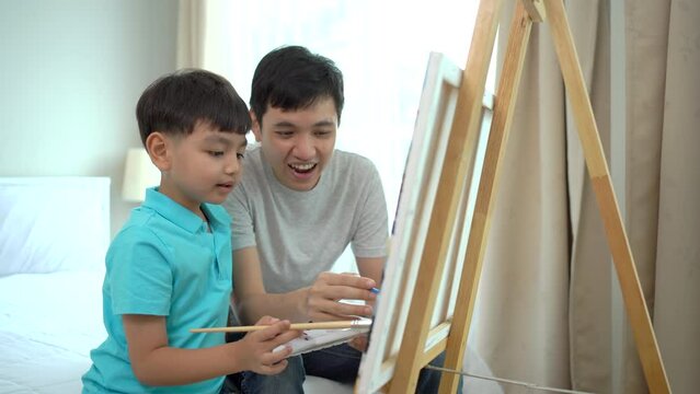 Happy Asian little boy enjoy drawing and painting a water color on a canvas with his father in the free time at home. Father teaching his son to draw and paint a picture. Creativity skill development.