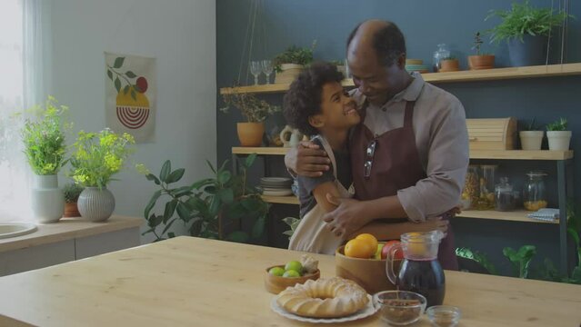 Happy little African American boy and grandfather smiling, hugging each other and posing for camera by kitchen table with homemade Meskouta cake and Zobo tea