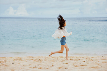 Sports woman runs along the beach in summer clothes on the sand in a yellow T-shirt and denim shorts white shirt flying hair ocean view