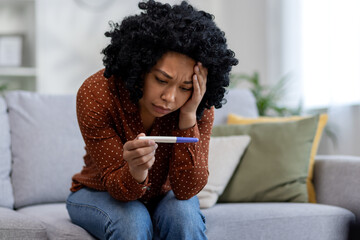 Frustrated and sad young african american woman crying sitting on sofa at home and holding negative pregnancy test